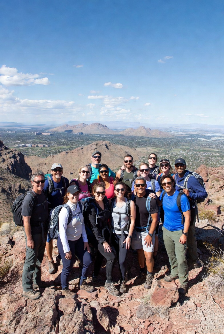 A group of about fifteen people at the summit of a Phoenix-area peak, Valley visible behind, blue sky