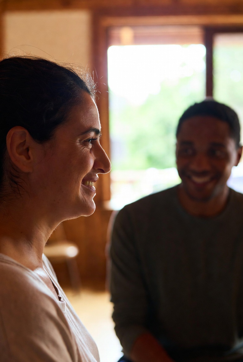Two adults in conversation in a warm clinical room, profile foreground, natural light from a window with greenery behind